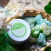 Detailed product shot of Sweet Clover CBD Body Cream in a white jar with green label, positioned beside sugar-coated green CBD & THC gummies, decorated with small white flowers, green leaves, and lace on a natural background.