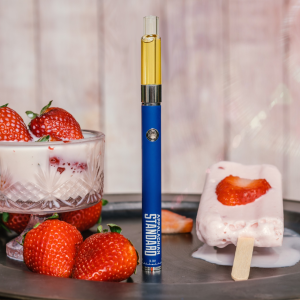 A product photography setup featuring a blue Appalachian Standard vape pen from Asheville, NC, labeled with 'APPALACHIAN STANDARD' and '3.3V' standing upright on a dark round tray. The pen contains calming plant goodness. Around it are fresh strawberries, a crystal dessert cup containing what appears to be strawberry yogurt or ice cream topped with two strawberries, and a strawberry popsicle or ice cream bar on a wooden stick melting slightly. The background is a soft pink-white color.