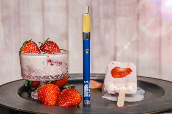 A product photography setup featuring a blue Appalachian Standard vape pen from Asheville, NC, labeled with 'APPALACHIAN STANDARD' and '3.3V' standing upright on a dark round tray. The pen contains calming plant goodness. Around it are fresh strawberries, a crystal dessert cup containing what appears to be strawberry yogurt or ice cream topped with two strawberries, and a strawberry popsicle or ice cream bar on a wooden stick melting slightly. The background is a soft pink-white color.