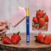 A styled product photo of a blue Appalachian Standard vape pen from Asheville, NC, labeled with 'APPALACHIAN STANDARD' and '3.3V' standing upright on a round wooden slice. The device offers calming plant goodness. It's surrounded by fresh red strawberries. In the blurred background, there's a crystal cup filled with more strawberries and a hand holding what appears to be a pink popsicle or ice cream bar. The hand has blue nail polish and is wearing rings. The background has a soft pink tone.
